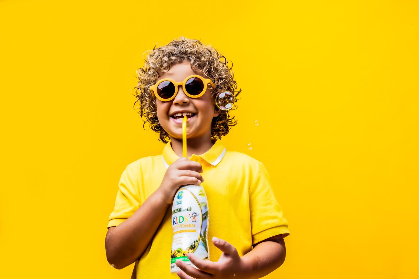 kid in yellow sunglasses and yellow shirt smiling and drinking from a drink against a yellow background